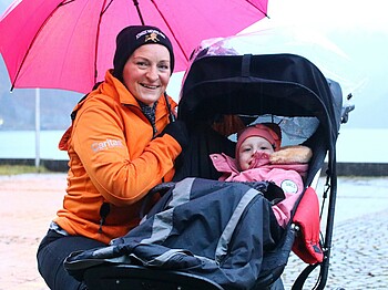 Eine Frau kniet mit Regenschrim bei einem Kind im Kinderwagen.