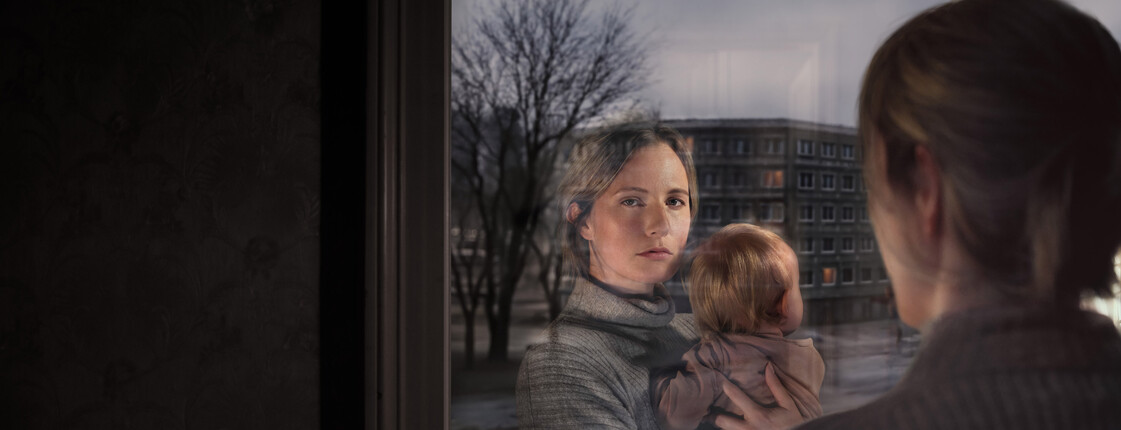 Eine Frau hält ein kleines Kind im Arm und schaut beim Fenster hinaus. In der Scheibe sieht man ihre Reflexion.