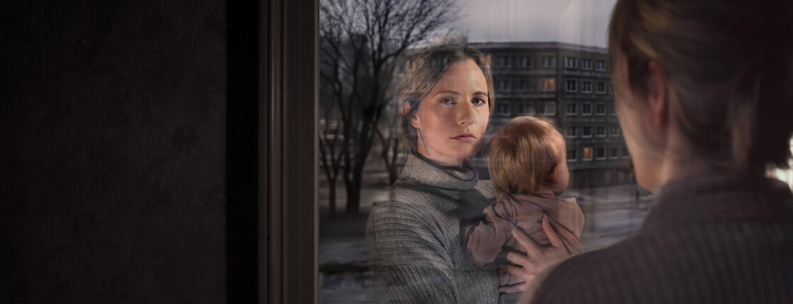Eine Frau hält ein kleines Kind im Arm und schaut beim Fenster hinaus. In der Scheibe sieht man ihre Reflexion.
