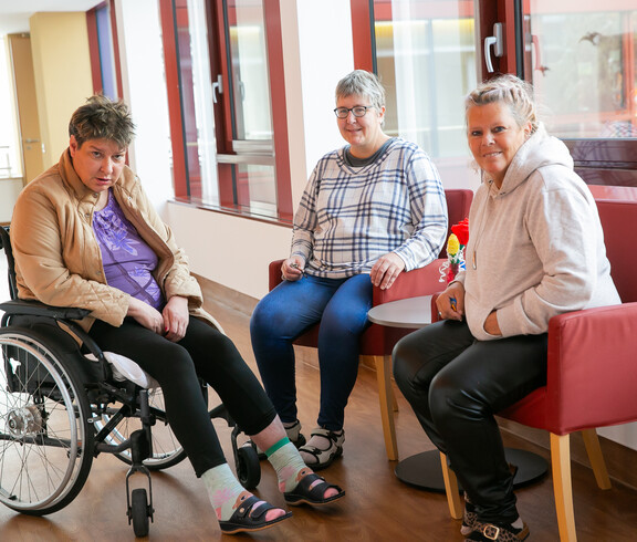 Drei Frauen sitezen zusammen und plaudern - links eine Frau im Rollstuhl, die anderen beiden eher rechst sitzen auf roten Stühlen.