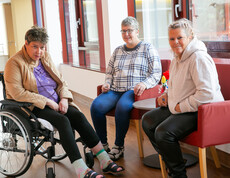 Drei Frauen sitezen zusammen und plaudern - links eine Frau im Rollstuhl, die anderen beiden eher rechst sitzen auf roten Stühlen.