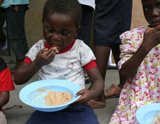 Ein Kind isst mit den Fingern nahrhaften Brei aus einem blauen Teller, neben ihm sitzt ein weiteres Kind mit Essen in der Hand