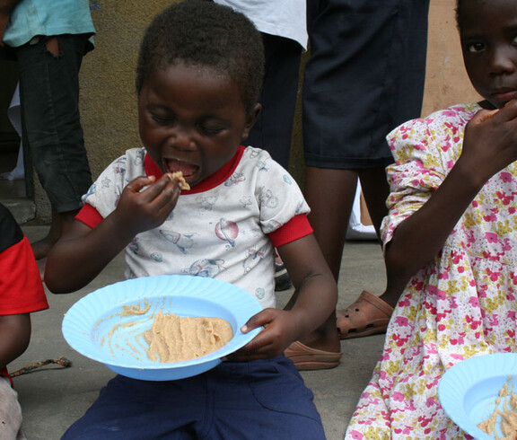 Ein Kind isst mit den Fingern nahrhaften Brei aus einem blauen Teller, neben ihm sitzt ein weiteres Kind mit Essen in der Hand