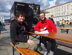 Zwei Personen sitzen auf Bänken auf einem öffentlichen Platz und schauen in die Kamera. 