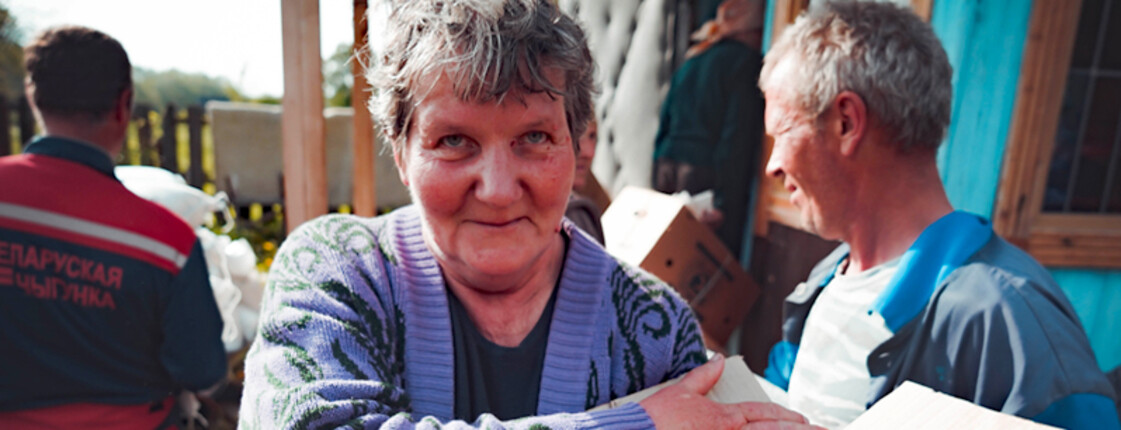 Ältere Frau hält ein Paket in den Armen, während im Hintergrund weitere Personen mit Kartons vor einem Haus stehen.