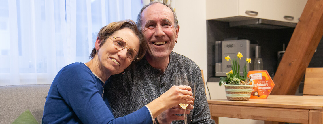 Frau lehnt Kopf an die Schulter von einem Mann, beide halten ein Glas Sekt in der Hand und lächeln in die Kamera