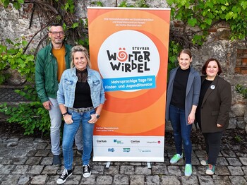 Vier Erwachsene stehen um ein oranges Plakat von der Veranstaltung Wörterwirbel