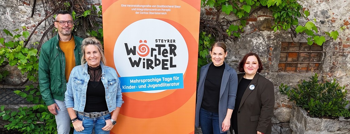 Vier Erwachsene stehen um ein oranges Plakat von der Veranstaltung Wörterwirbel
