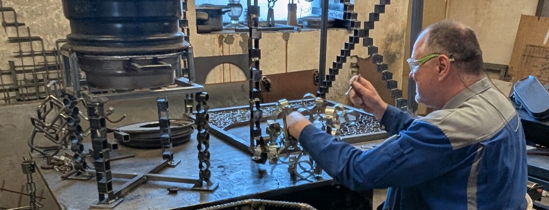 Mann von hinten rechts im Bild sitz am Tisch und arbeitet mit Metallteilen.