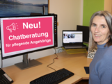 Eine Frau sitzt am Schreibstisch und lächelt in die Kamera. Am Bildschrim des Computers ist zu lesen: Neu Chatberatung für Pflegende Angehörige
