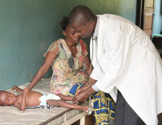 Ein Arzt untersucht ein krankes Kleinkind, das weinend auf einer Liege neben seiner Mutter liegt