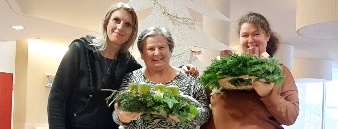Drei Frauen schauen in die Kamera, die beiden rechts und in der Mitte halten je einen Adventkranz in die Höhe.