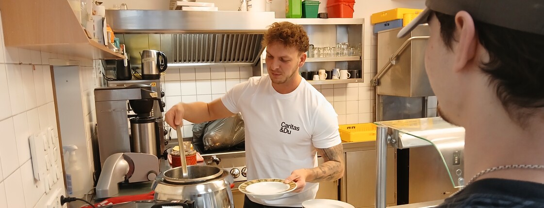 Ein Mann schöpft Suppe aus einem großen Topf in einen Suppenteller