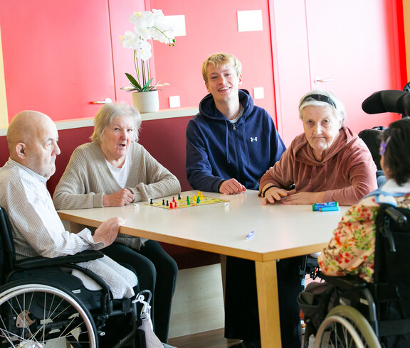 Zwei Personen im Rollstuhl und drei weitere Personen sitzen gemeinsam an einem Tisch.