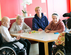 Zwei Personen im Rollstuhl und drei weitere Personen sitzen gemeinsam an einem Tisch.