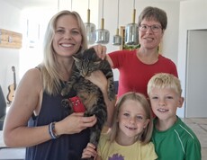 Eine blonde Frau links im Bild hält eine Katze im Arm, zwei Kinder rechts daneben und dahinter eine weitere Frau, die die Katze streichelt.