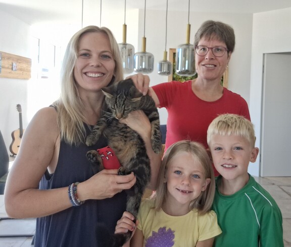 Eine blonde Frau links im Bild hält eine Katze im Arm, zwei Kinder rechts daneben und dahinter eine weitere Frau, die die Katze streichelt.