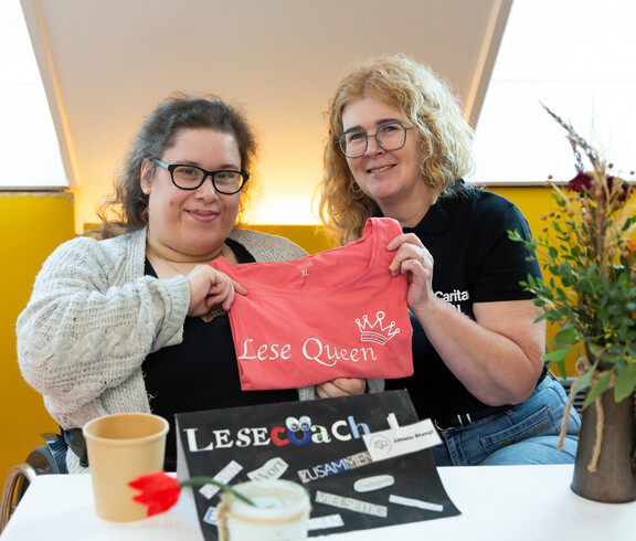 Zwei Frauen halten ein T-Shirt, auf dem Lese Queen drauf steht, in die Kamera. Vor ihnen auf dem Tisch steht ein Schild, auf dem Lesecoach steht.