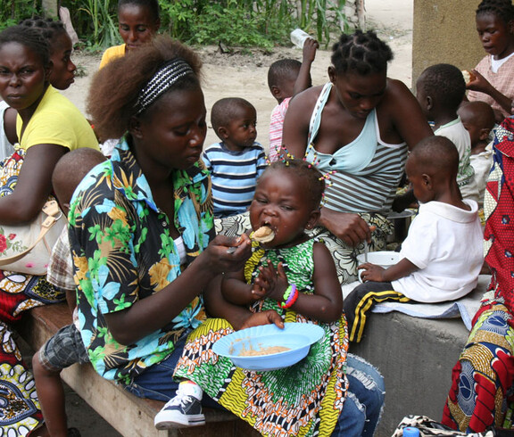 Mehrere Mütter sitzen mit ihren Kindern im Freien, während eine Mutter ihr Kind mit nahrhaftem Brei füttert. Im Hintergrund essen weitere Kinder