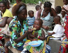 Mehrere Mütter sitzen mit ihren Kindern im Freien, während eine Mutter ihr Kind mit nahrhaftem Brei füttert. Im Hintergrund essen weitere Kinder