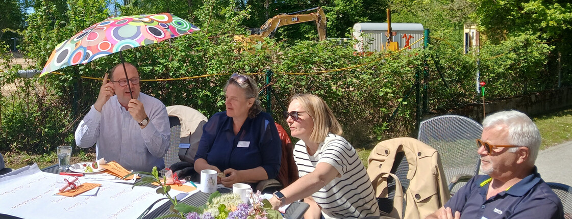 4 Personen sitzen an einem Gartentisch. Ein Herr hält einen bunten Regenschirm über seinen Kopf.