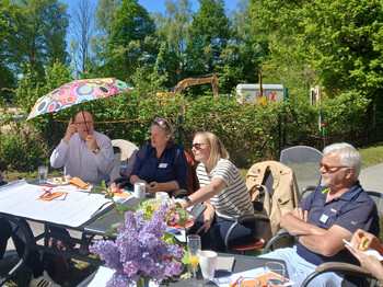 4 Personen sitzen an einem Gartentisch. Ein Herr hält einen bunten Regenschirm über seinen Kopf.
