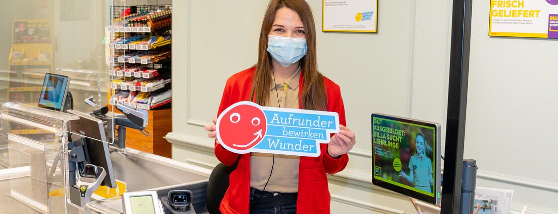 Eine Frau steht hinter der Supermarkt-Kassa, sie hält ein Schild in der Hand mit dem Schriftzug "Aufrunder bewirken Wunder".