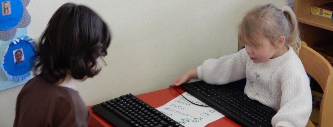 Zwei Kinder sietzen bei einem roten Tisch. Darauf liegen zwei Tastaturen.