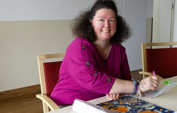 Eine Frau mit dunklen Haaren und lila T-Shirt sitzt an einem Tisch und malt ein Bild
