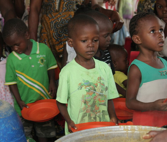 Mehrere Kinder stehen in einer Schlange mit leeren orangefarbenen Tellern in der Hand und warten auf eine Essensausgabe. Im Vordergrund ist ein großer Topf mit Brei oder Suppe zu sehen, aus dem gerade geschöpft wird. Die Kinder blicken ernst oder geduldig nach vorne, im Hintergrund sind weitere Kinder und Erwachsene zu erkennen
