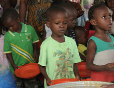 Mehrere Kinder stehen in einer Schlange mit leeren orangefarbenen Tellern in der Hand und warten auf eine Essensausgabe. Im Vordergrund ist ein großer Topf mit Brei oder Suppe zu sehen, aus dem gerade geschöpft wird. Die Kinder blicken ernst oder geduldig nach vorne, im Hintergrund sind weitere Kinder und Erwachsene zu erkennen