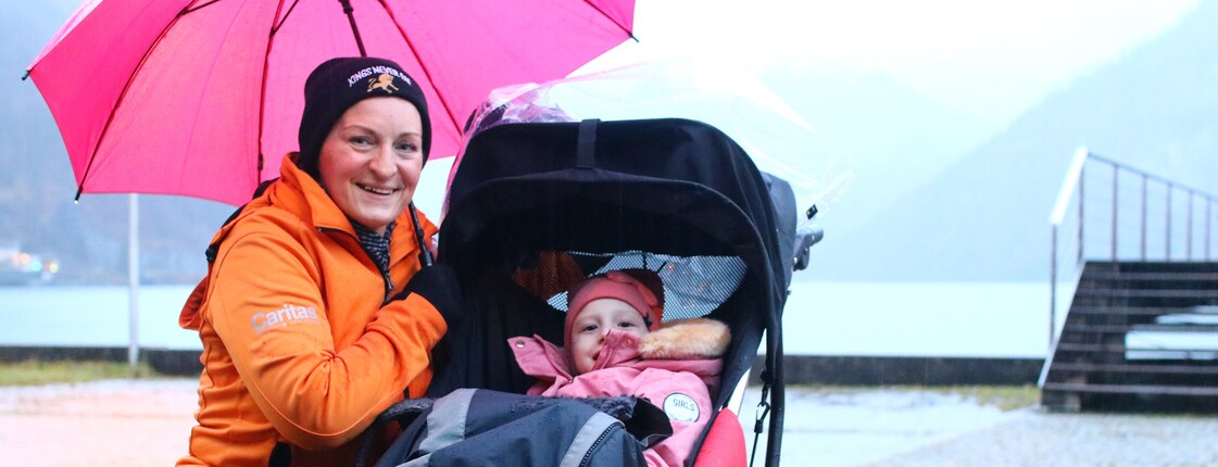 Eine Frau kniet mit Regenschrim bei einem Kind im Kinderwagen.