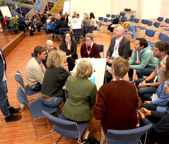 Blick auf eine Gruppe von Personen, die rund um einen Tisch sitzen und sich unterhalten.