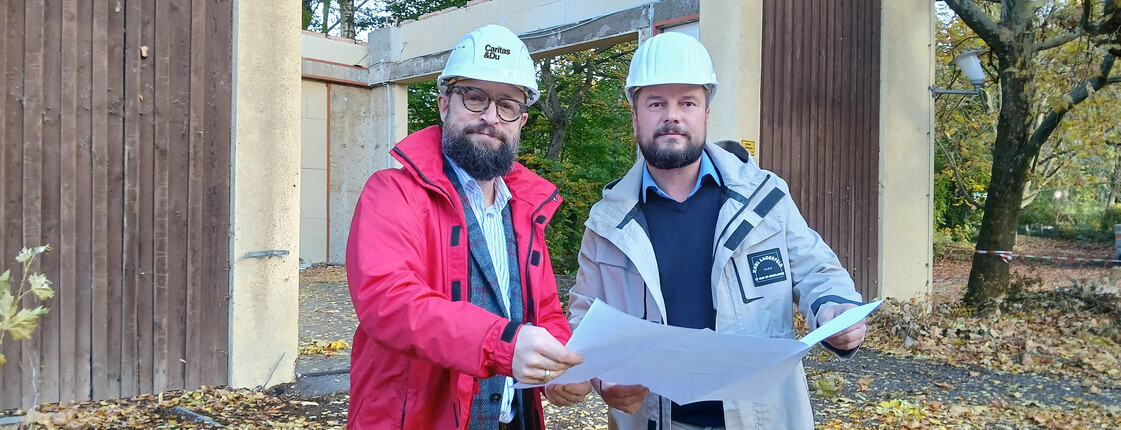 Zwei Männer mit Bauhelm stehen vor einem Gebäude, das bereits zum Teil abgetragen wurde.