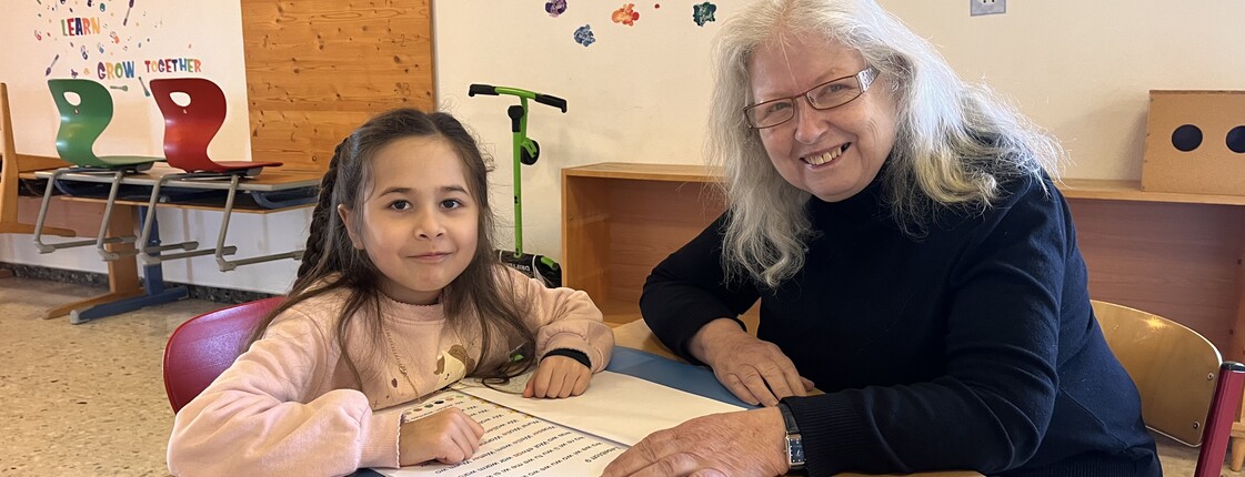 Ein Mädchen mit langen dunklen Haaren im Volksschulalter mit einer älteren Dame mit langen weißen Haaren sitzen beim Tisch mit einer Lesemappe. Beide schauen in die Kamera.