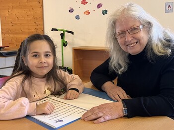 Ein Mädchen mit langen dunklen Haaren im Volksschulalter mit einer älteren Dame mit langen weißen Haaren sitzen beim Tisch mit einer Lesemappe. Beide schauen in die Kamera.