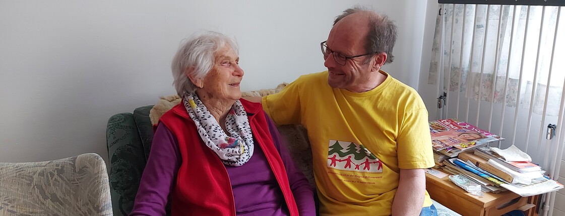 Eine ältere Dame, links im Bild, und ein Herr rechts im Bild sitzen auf einem Sofa und unterhalten sich.