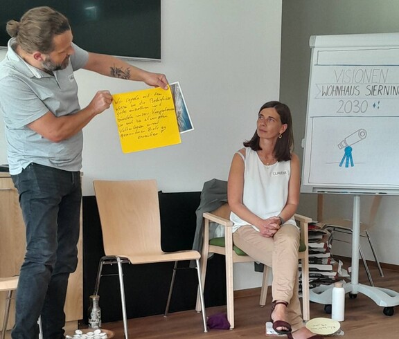 Frau sitzt auf Stuhl vor einem Flipchart, Mann hält eine gelbe Tafel zur Kamera hin.