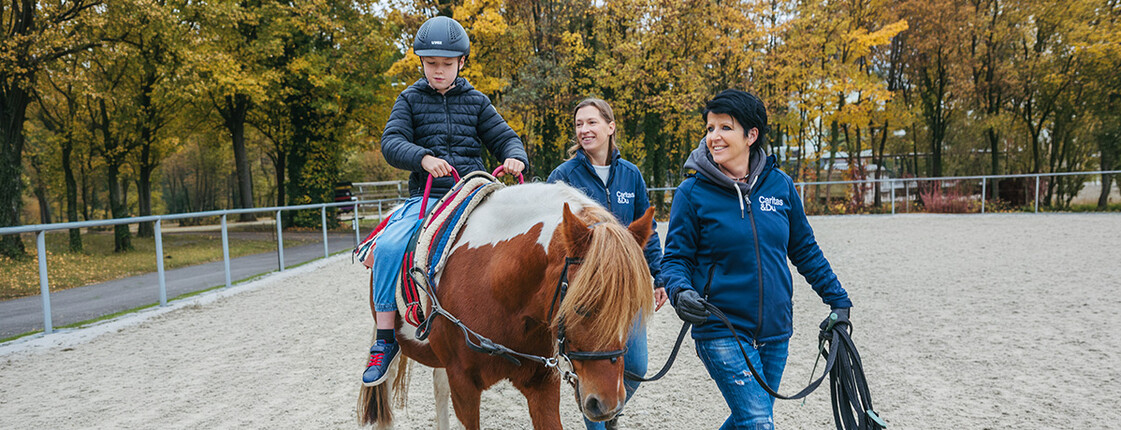 Ein Kind sitzt auf einem Pferd und wird im Freien von zwei Personen begleitet.