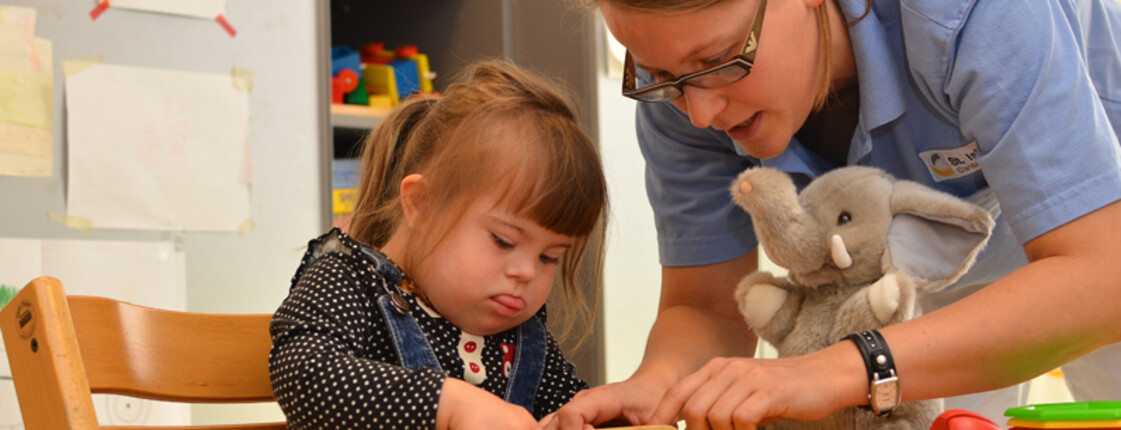 Ein kleines Mädchen sitzt mit einer Therapeutin an einem Tisch und spielt mit Holzspielzeug.