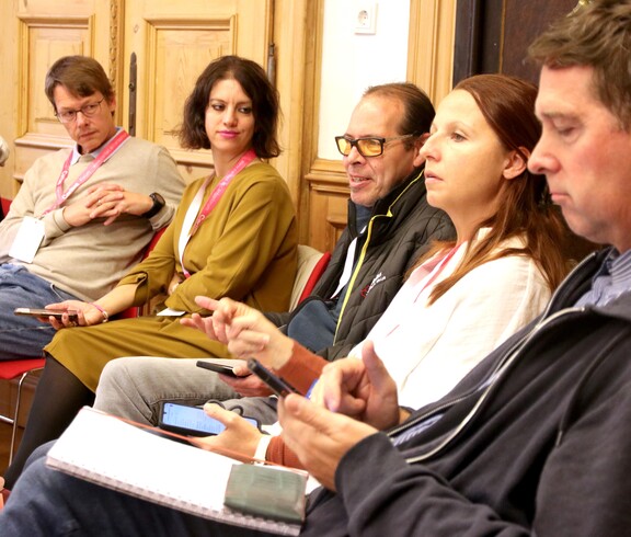 Fünf Personen sitzen auf Sesseln und hören den Vortragenden zu.