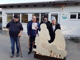Krippenfiguren aus Holz sind auf einem kleinen Anhhänger geladen. Ein paar Männer stehen dahinter und schauen in die Kamera.
