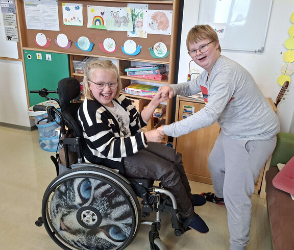 Ein Mädchen im Rollstuhl hält die Hände von einem lächelnden Mädchen