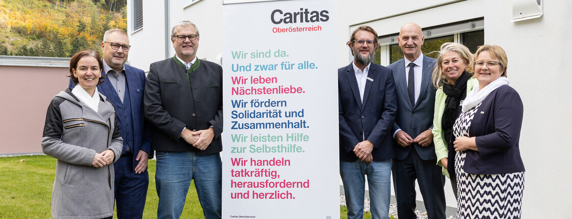 Sieben Personen stehen vor einem Gebäude im Freien und rund um ein Caritas Roll-Up beisammen.