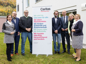 Sieben Personen stehen vor einem Gebäude im Freien und rund um ein Caritas Roll-Up beisammen.