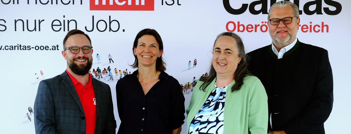 Vorstand der Caritas Oberösterreich Vier Personen stehen vor einem Caritas-Plakat und lächeln.
