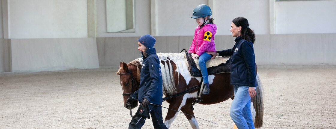 Ein Kind sitzt auf einem Pferd in einer Halle und wird von zwei Personen begleitet.