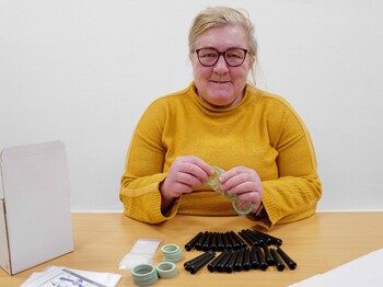 Frau in gelbem Pullover verpakct Kleinteile am Tisch.