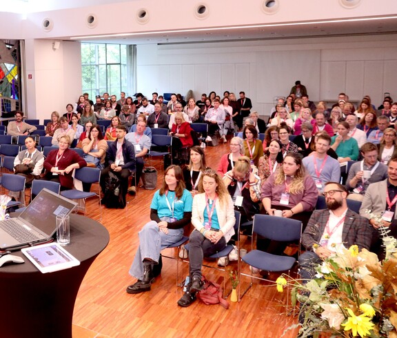 Ein Blick in einem vollgefüllten Saal, wo die Menschen auf Stühlen sitzen.