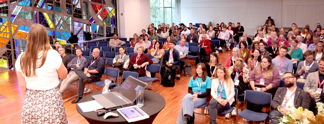 Ein Blick in einem vollgefüllten Saal, wo die Menschen auf Stühlen sitzen.